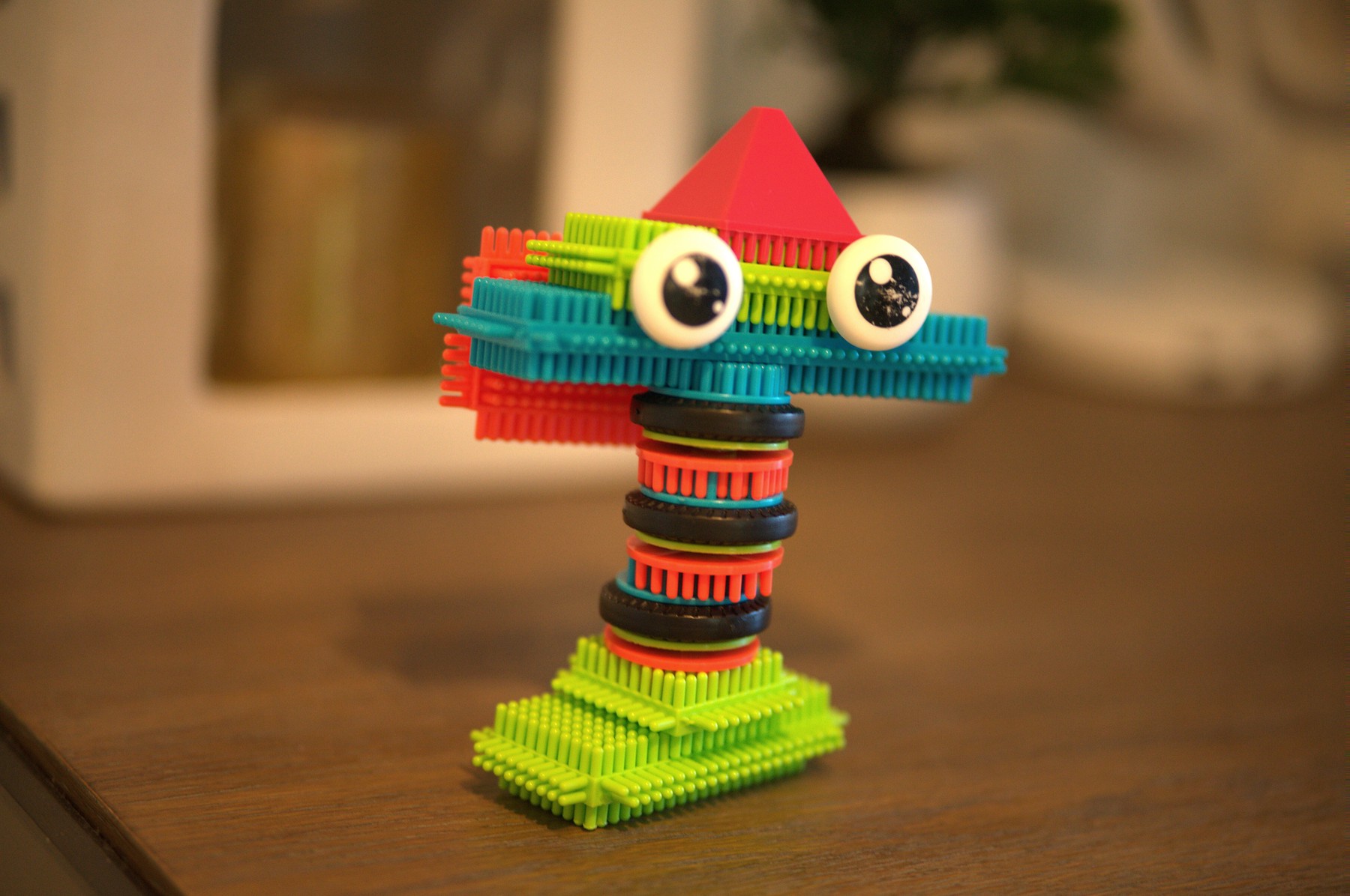 A creature built from bristle blocks standing on a wooden table — ring-stacked neck, wide teal head, two uneven googly eyes, and a red pyramid hat