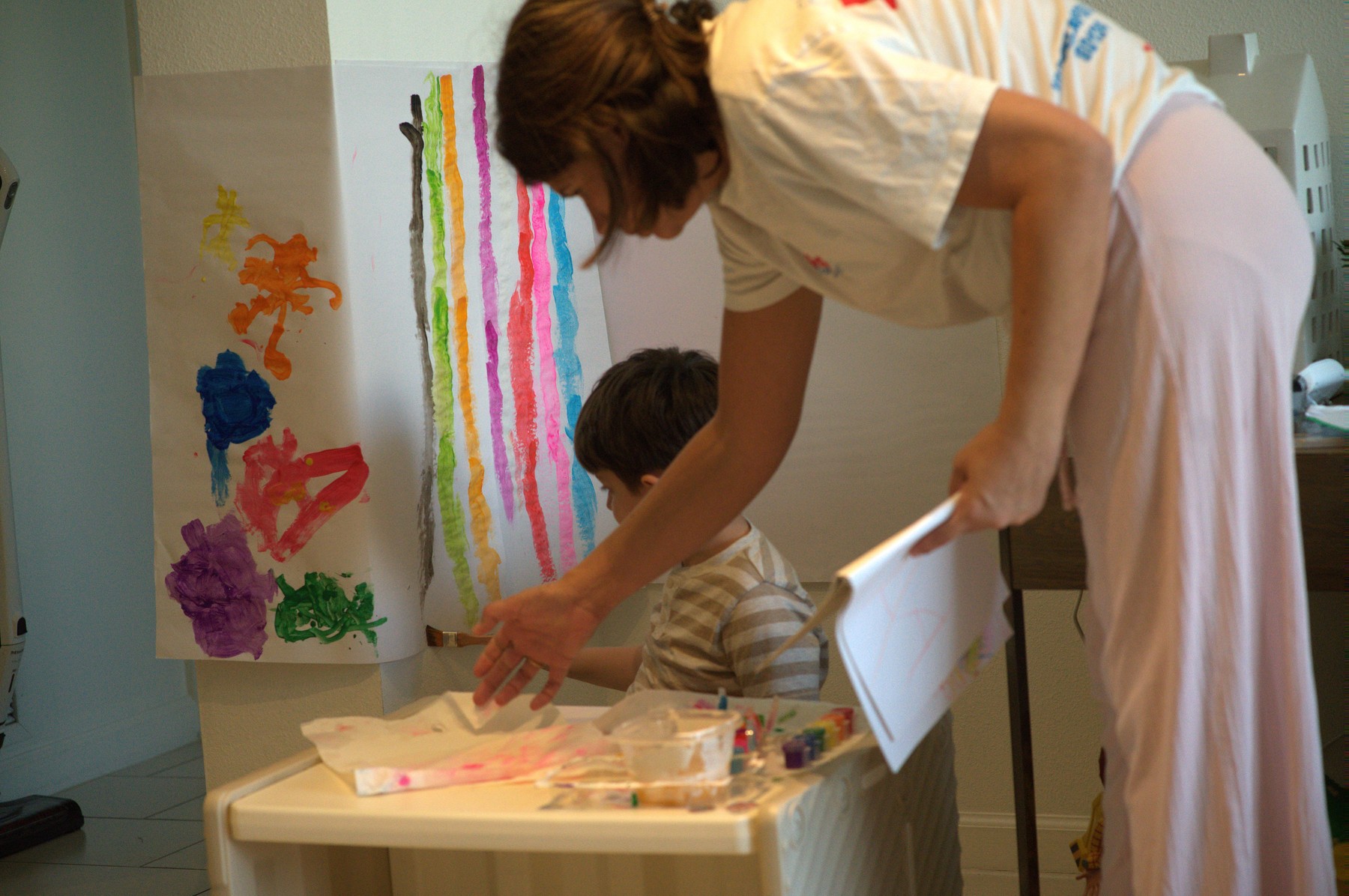 Lucas painting at a low table beneath two big wall sheets — rainbow stripes on one, bold blobby shapes on the other — while Lorena leans in beside him holding his sketchbook