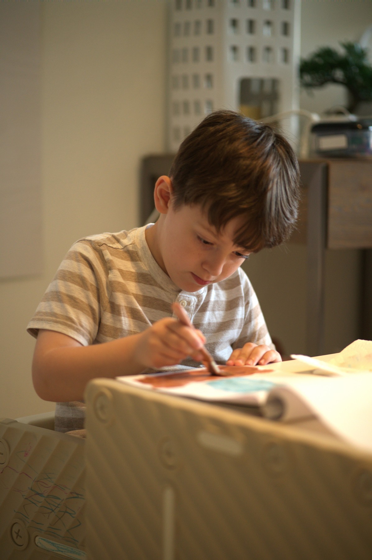 Lucas leaning over his paper with his head bowed, tongue tip showing, working a wide brush over rust-red and teal shapes