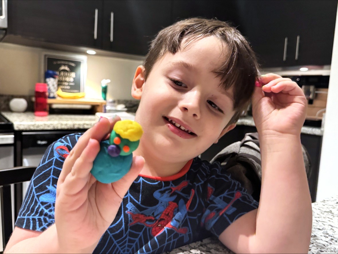 Lucas in the kitchen in a Spider-Man t-shirt, head tilted and smiling, holding a small play-doh figure up close to the camera in one hand — teal at the base, green above it, yellow on top — with a small pink piece pinched in his other hand beside his head