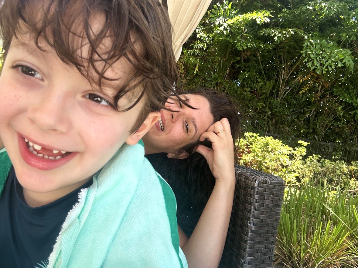 A close selfie at the pool: Lucas's face fills the left of the frame, his wet hair in damp curls over his forehead and a pale green towel over his shoulder, grinning into the camera while Lorena peeks in smiling behind him at the frame's edge