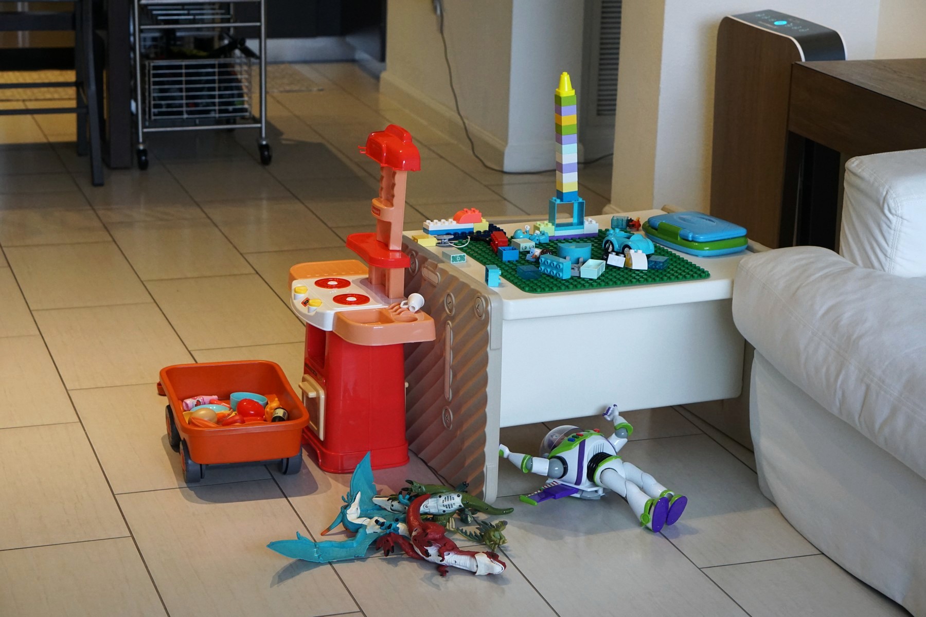 Lucas's empty play corner — a tall rainbow brick tower still standing on the white table, the red toy kitchen and a wagon of play food beside it, dinosaurs and a blue dragon heaped on the floor
