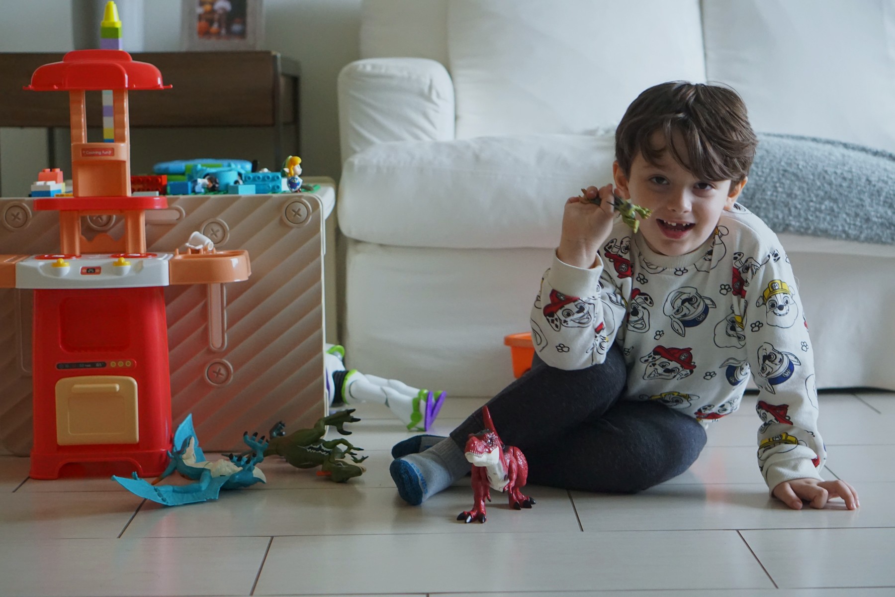 Lucas sitting cross-legged in his play corner, grinning at the camera, holding a small green dinosaur up to his ear like a telephone, a red T. rex standing in front of him