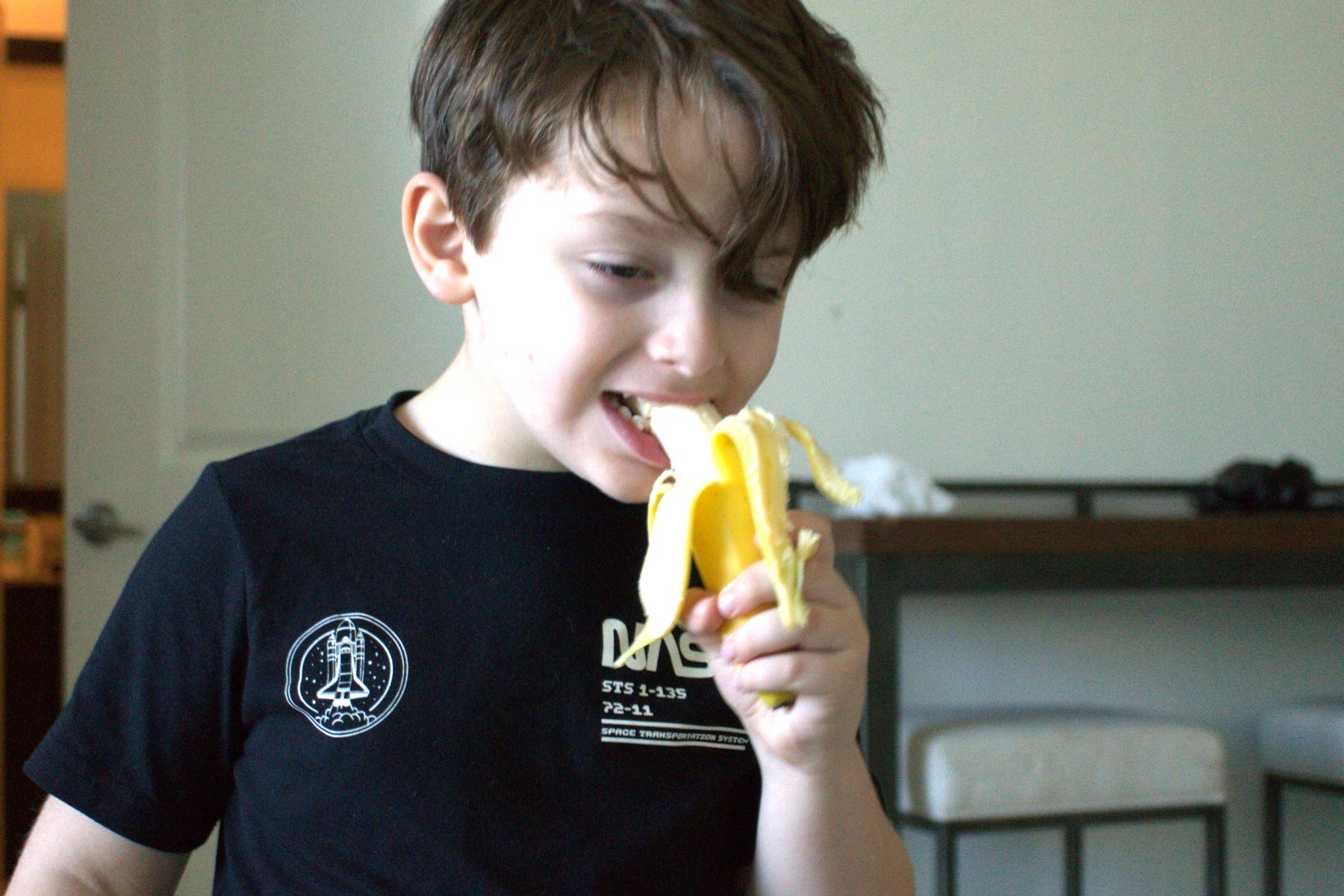 Lucas mid-bite into a peeled banana, eyes half closed, wearing a black T-shirt with a white Space Shuttle on it