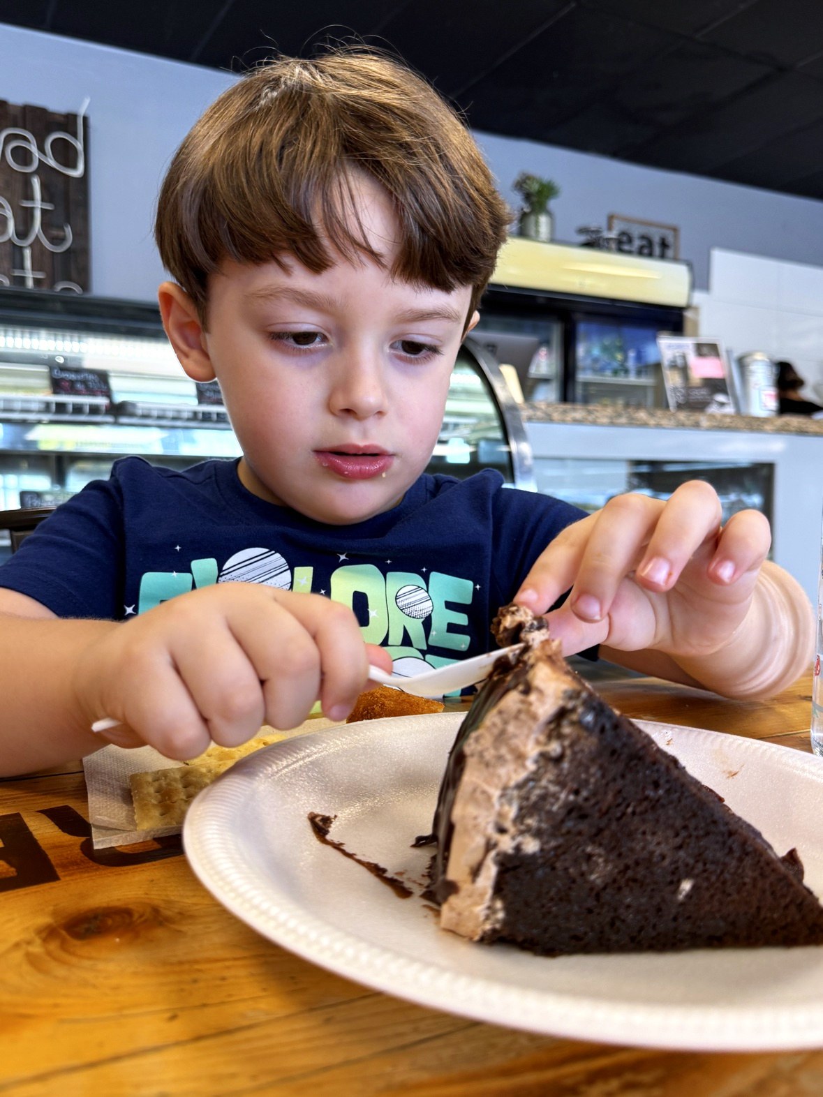 Lucas at a bakery counter cutting into a tall slice of chocolate cake on a white plate with a plastic spoon, the fingers of his other hand steadying the top of the slice, wearing a shirt that says EXPLORE