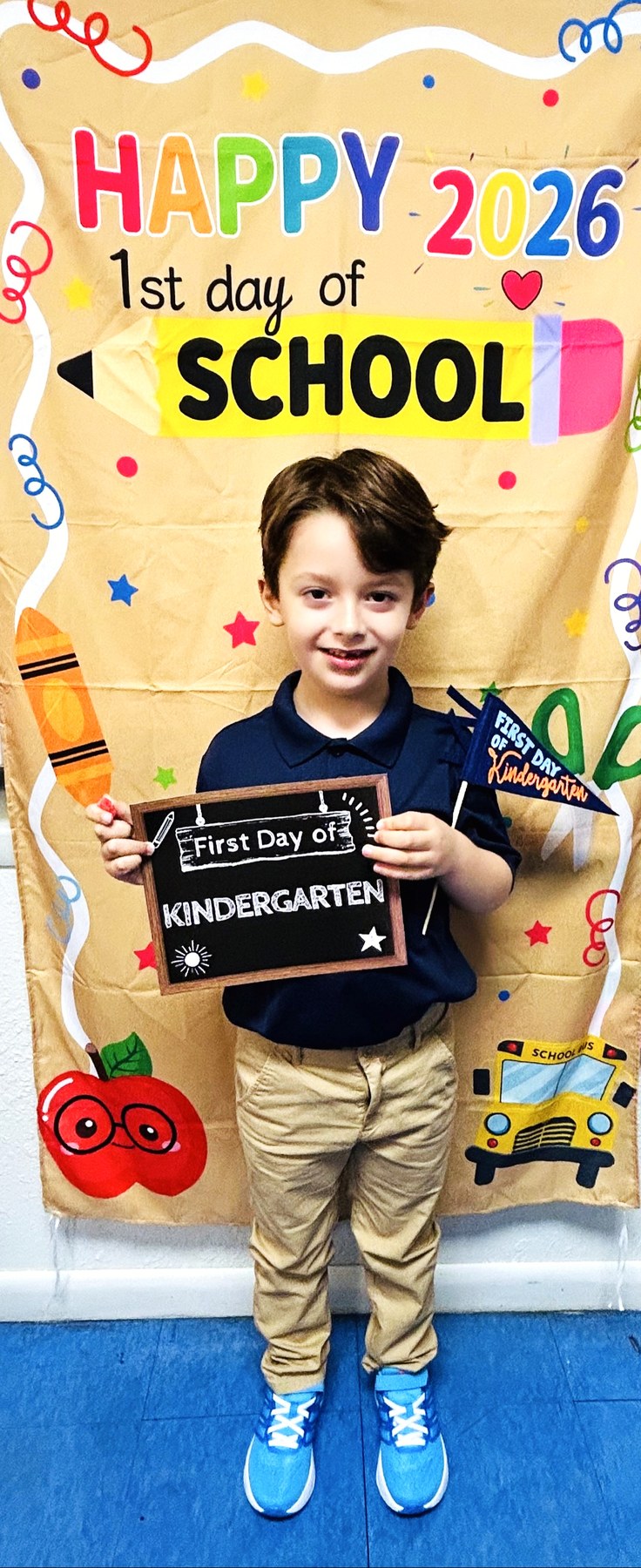 Lucas standing full-length in front of a printed backdrop that reads HAPPY 2026 and 1st day of SCHOOL, holding a framed board that reads First Day of KINDERGARTEN in both hands, wearing a navy polo shirt, khaki trousers and bright blue sneakers, smiling at the camera