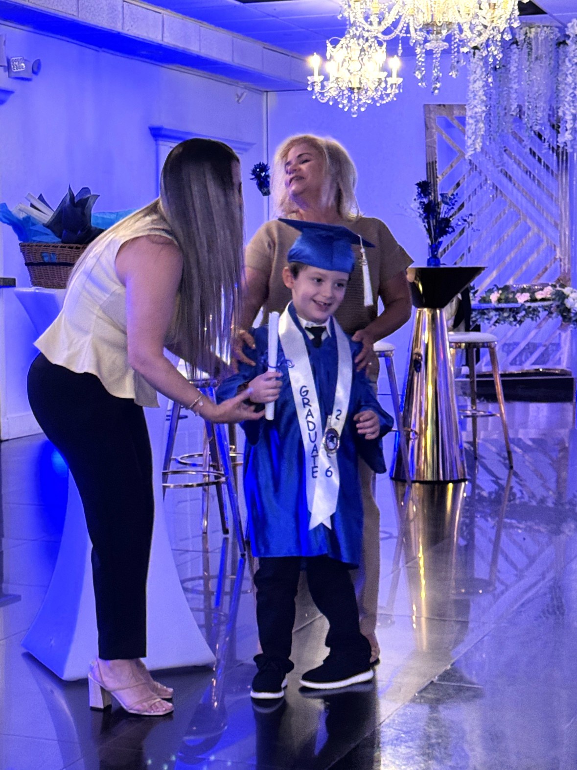 Lucas standing in the middle of a blue-lit ballroom in a blue cap and gown, a white sash reading “GRADUATE” across his chest, a medal at his chest, and a rolled diploma in his hand — a woman bent toward him at his side, another woman standing behind him — smiling
